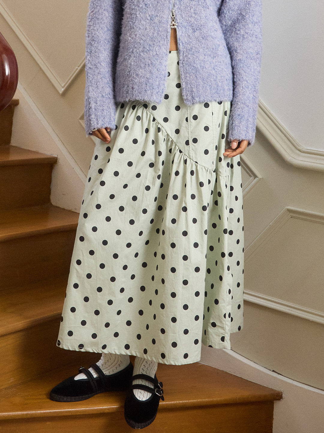 Person wearing a polka dot skirt and light purple sweater on wooden stairs.